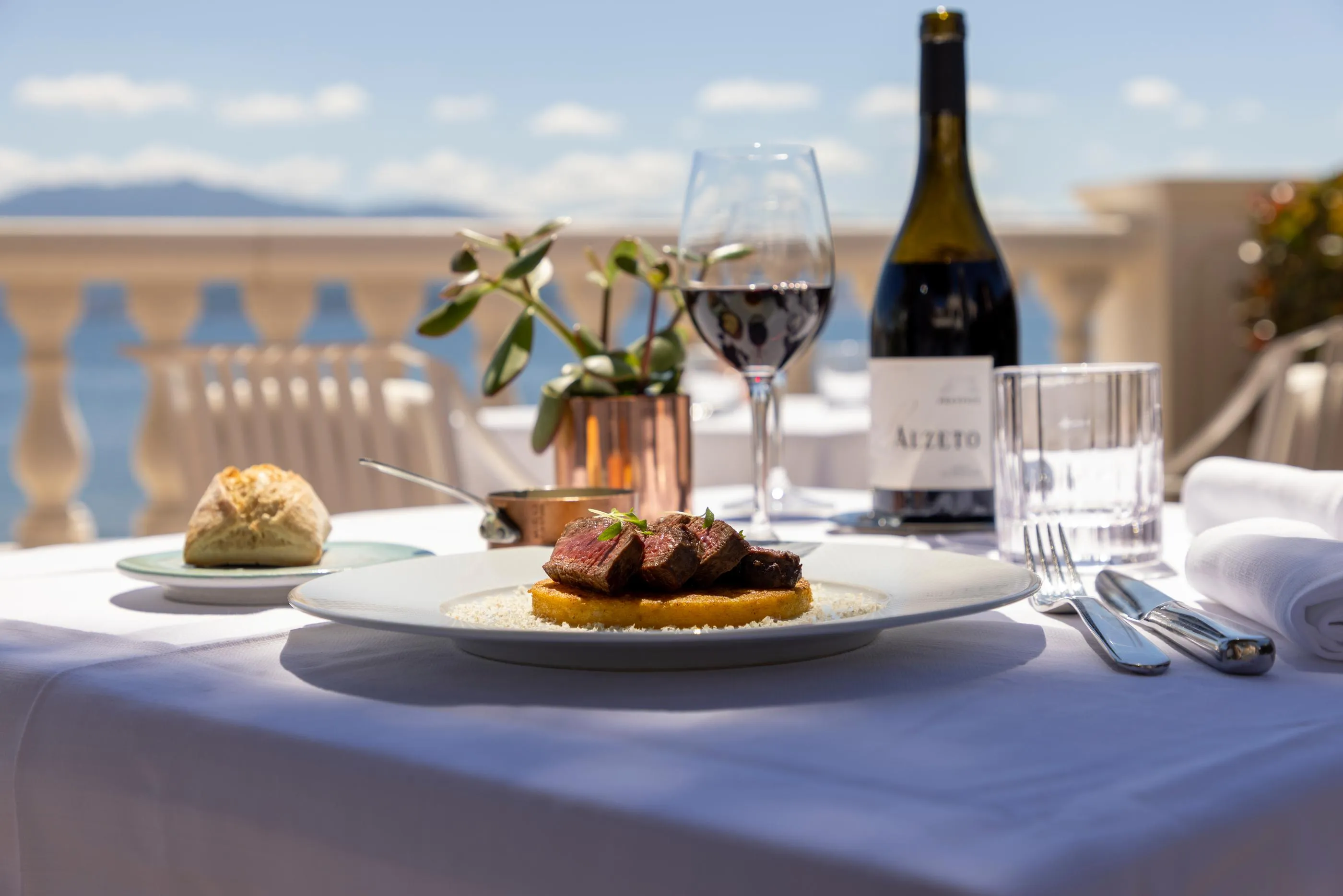 Table dressée avec un plat de viande, un verre de vin, une bouteille de vin et des couverts, en extérieur avec vue sur un balcon