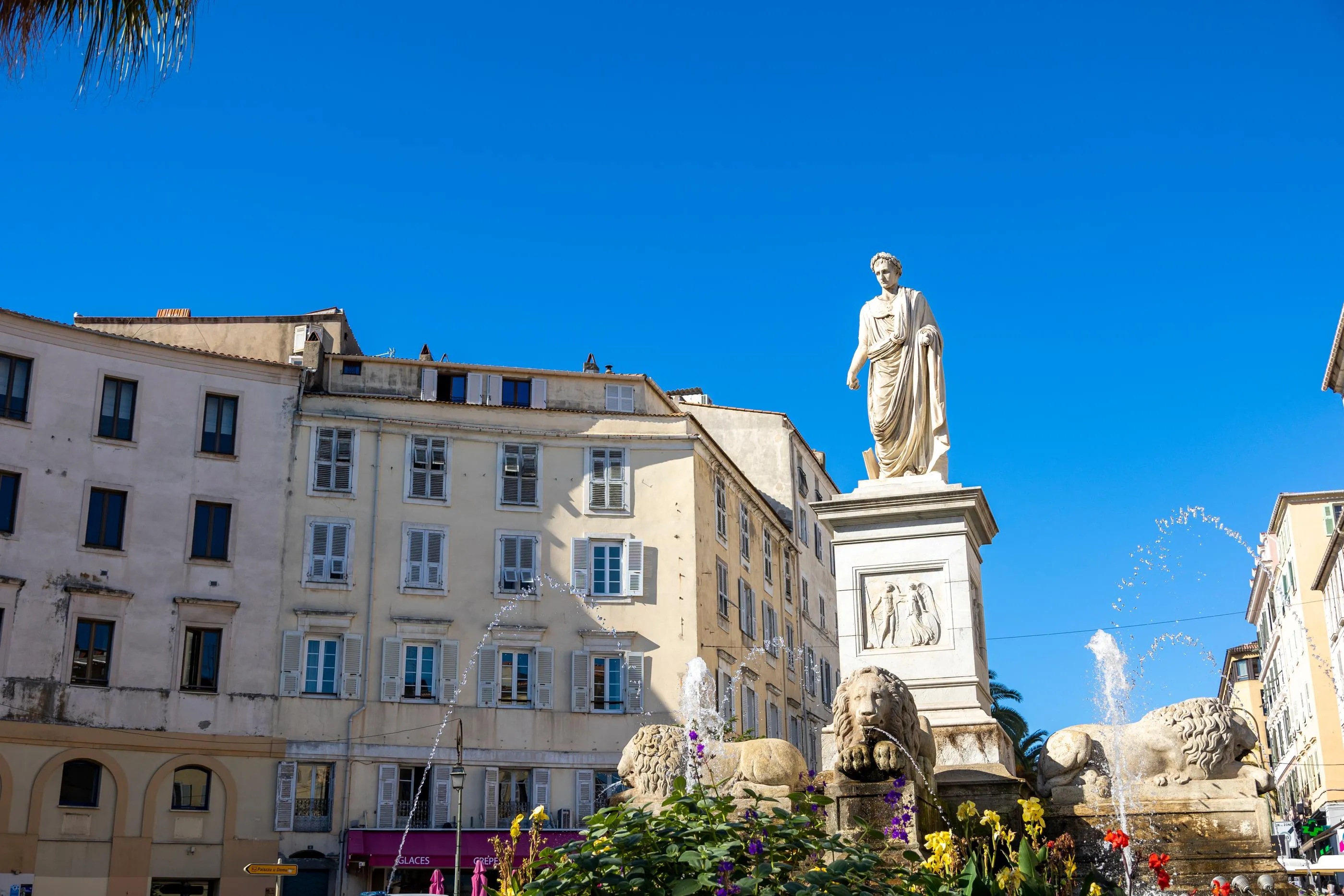 Statue d'une figure historique sur un piédestal orné, entourée de jets d'eau, devant des bâtiments anciens aux fenêtres à volets