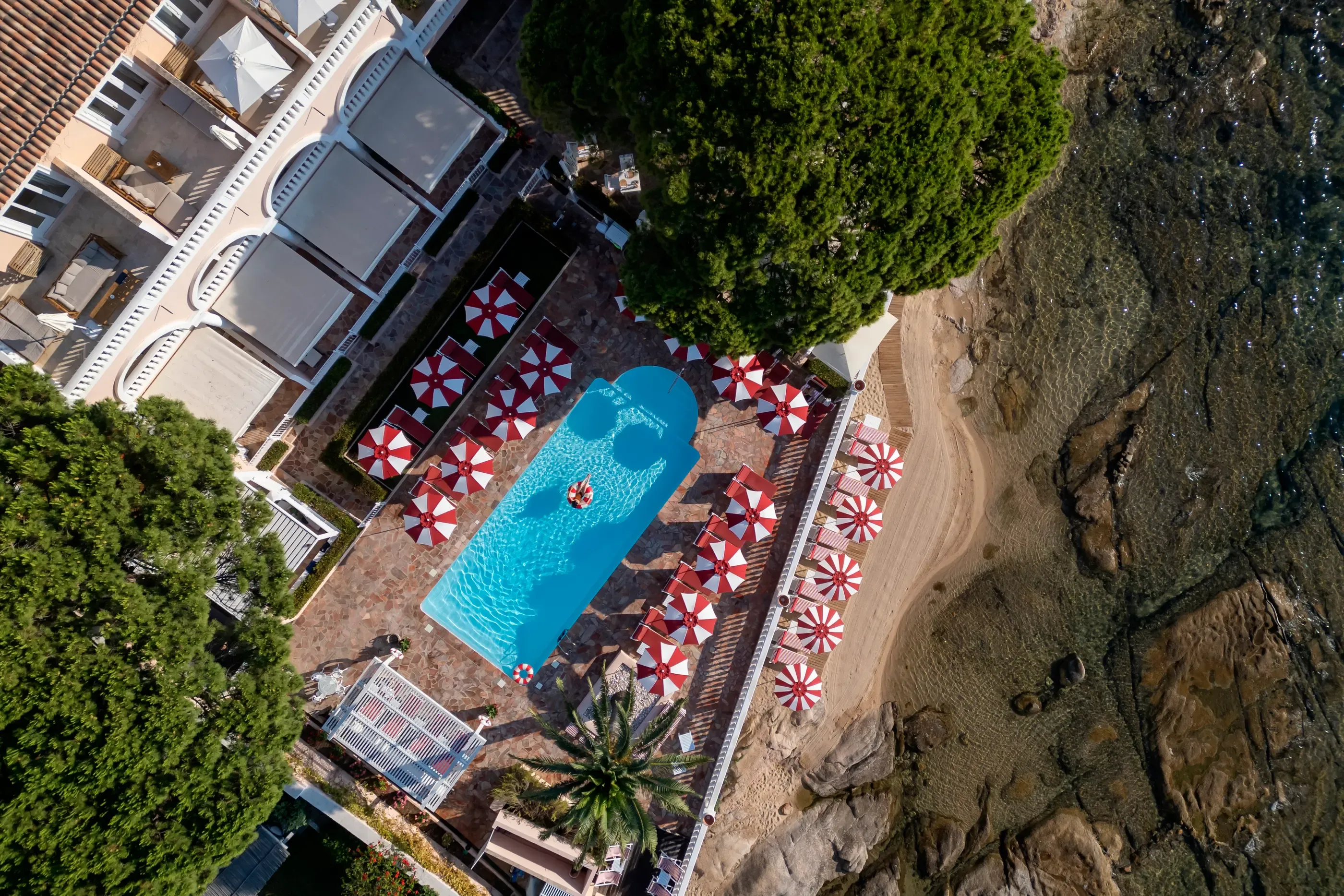 Vue aérienne d'une piscine entourée de parasols rouges et blancs, située près d'un bâtiment et d'une plage de sable